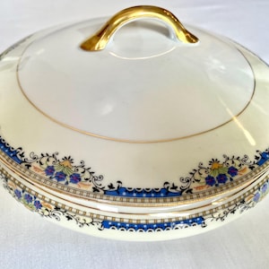 May include: A white and cream-colored porcelain serving dish with a lid. The dish features gold handles and a gold rim. A blue, black, and yellow floral pattern decorates the rim of the dish. The lid has a gold handle.