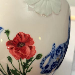 May include: A white ceramic teacup with a red poppy and blue floral design. The cup features a raised white flower and a blue rim. The poppy has green leaves and buds. The design is delicate and detailed.