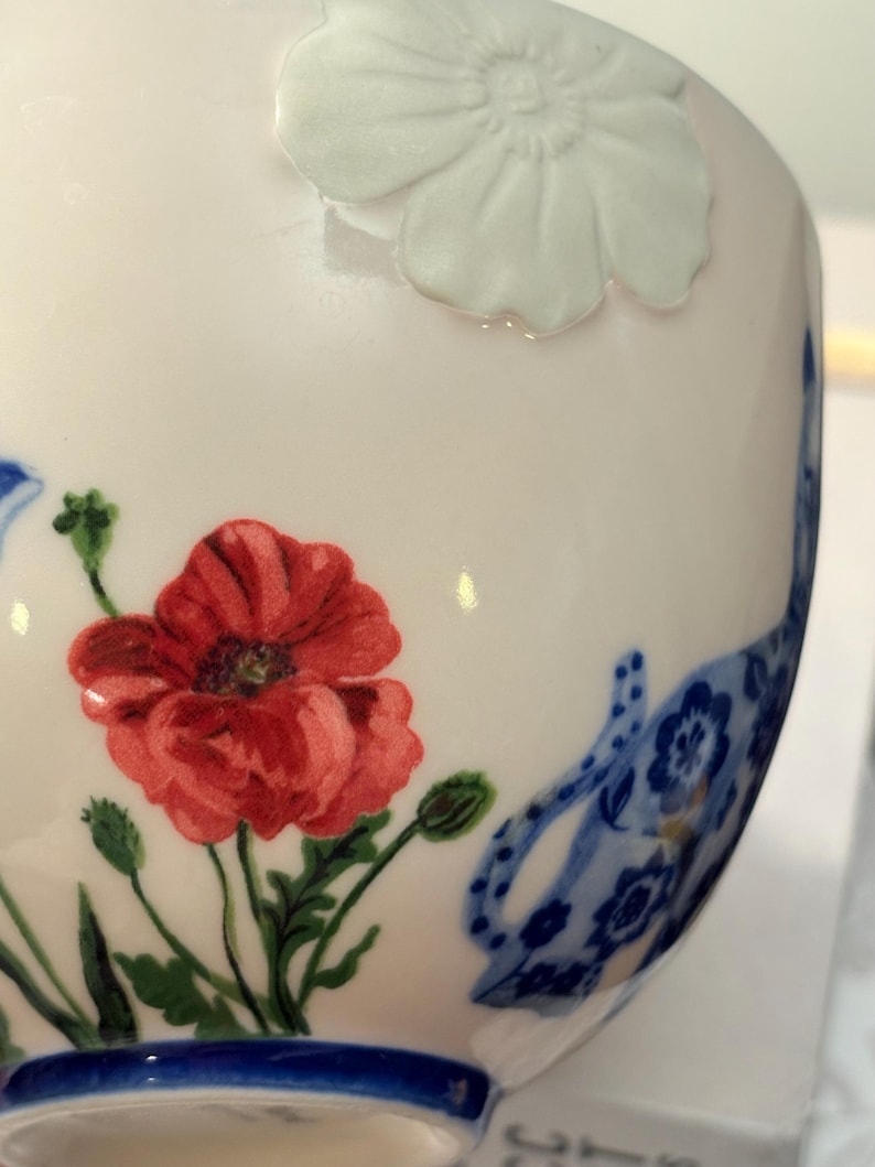 May include: A white ceramic teacup with a red poppy and blue floral design. The cup features a raised white flower and a blue rim. The poppy has green leaves and buds. The design is delicate and detailed.