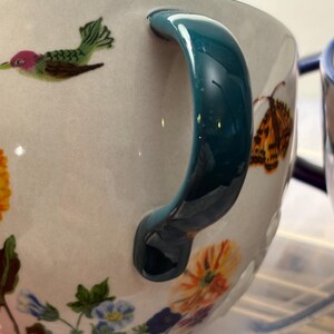 May include: Close-up of a ceramic mug with a teal handle and floral and hummingbird illustrations. The mug features a white background with colorful flowers and a hummingbird design. The handle is a glossy teal color.