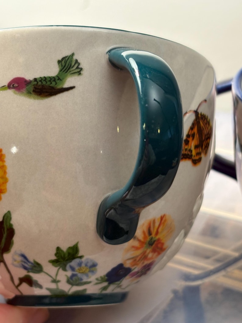 May include: Close-up of a ceramic mug with a teal handle and floral and hummingbird illustrations. The mug features a white background with colorful flowers and a hummingbird design. The handle is a glossy teal color.