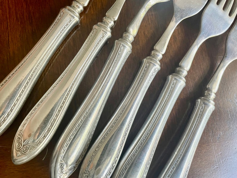 May include: Six silver forks with ornate handles. The forks have a decorative pattern on the handles and are arranged in a row.