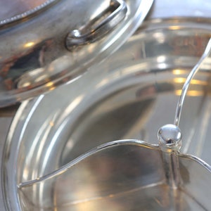 Puede incluir: Un plato de servir plateado con tapa y un divisor extraíble. El plato está hecho de metal pulido y tiene un borde decorativo.