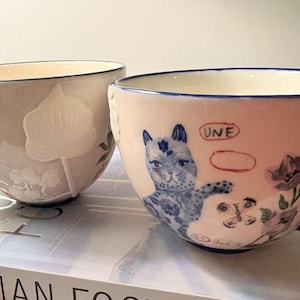 May include: Two ceramic teacups with handles. One is light gray with a butterfly and floral relief. The other is pink with a blue cat illustration, flowers, and the word "UNE". Both have blue accents.