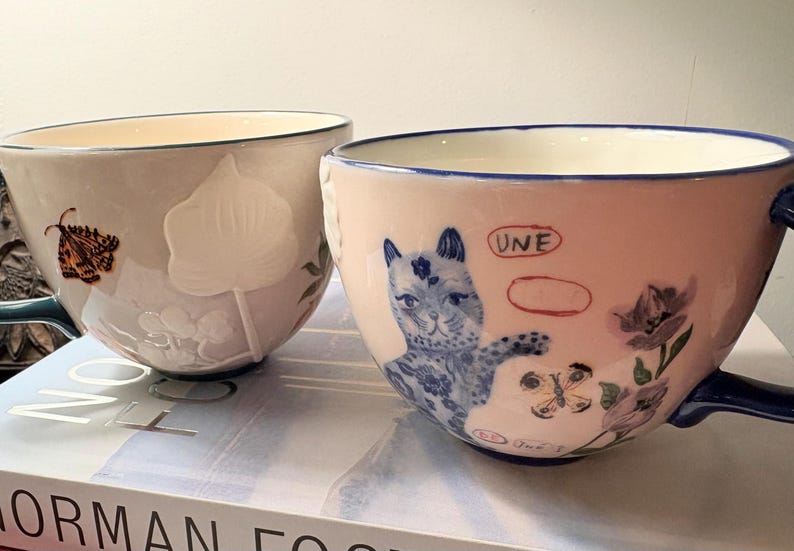 May include: Two ceramic teacups with handles. One is light gray with a butterfly and floral relief. The other is pink with a blue cat illustration, flowers, and the word "UNE". Both have blue accents.