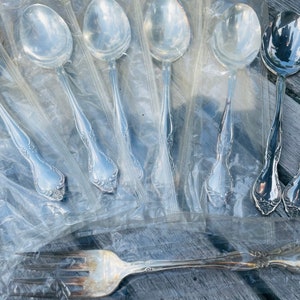 Könnte beinhalten: Ein Set aus acht silbernen Löffeln mit verzierten Griffen, verpackt in durchsichtigem Plastik. Die Löffel sind in einer Reihe angeordnet, mit den Griffen nach oben. Eine silberne Gabel ist ebenfalls auf dem Bild zu sehen.