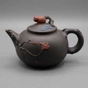 May include: A dark brown clay teapot featuring a sculpted branch with reddish-brown flower blossoms on the body. The lid includes a reddish-brown bud-shaped finial and a small blue leaf accent. The handle and spout are shaped like textured tree branches.