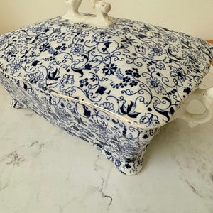 May include: A white and blue floral patterned serving dish with a lid. The dish has a rectangular shape with rounded corners, a gold trim, and a white handle on the lid. The dish is raised on small feet.