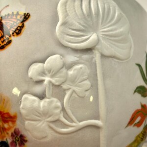 May include: A ceramic teacup with a light gray background. The cup features a raised white floral design and a painted butterfly with orange and black wings. Other painted flowers and greenery are also visible.