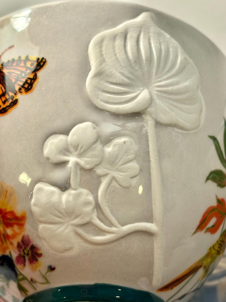 May include: A ceramic teacup with a light gray background. The cup features a raised white floral design and a painted butterfly with orange and black wings. Other painted flowers and greenery are also visible.