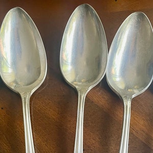 May include: Five silver spoons with a polished finish. The spoons are arranged in a row, with the bowls facing the viewer. The spoons are all the same size and shape.