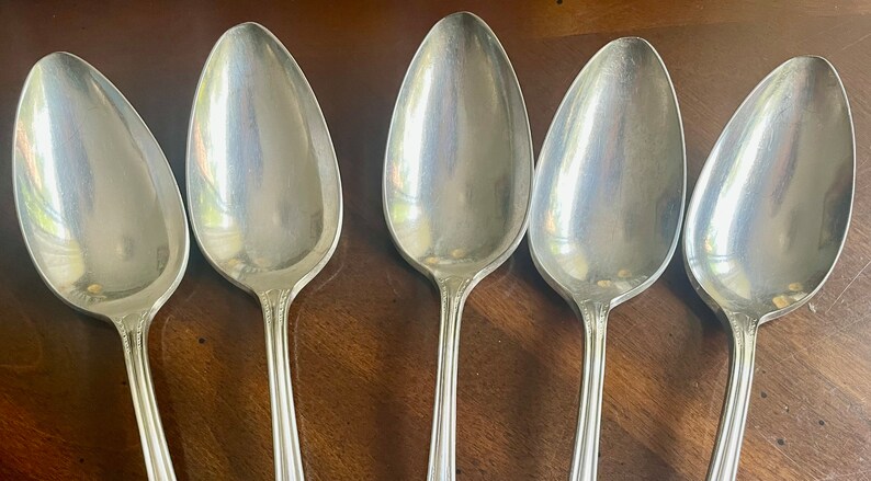 May include: Five silver spoons with a polished finish. The spoons are arranged in a row, with the bowls facing the viewer. The spoons are all the same size and shape.