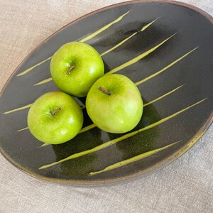 May include: A black oval ceramic platter with three green apples. The platter has yellow diagonal lines and a brown rim. The apples are Granny Smith apples.