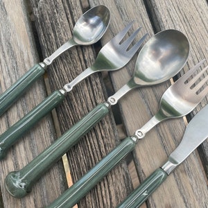 Puede incluir: Un juego de cuatro cubiertos plateados con mangos verdes. El juego incluye un tenedor, un cuchillo, una cuchara y una cuchara más pequeña.