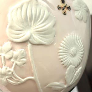 May include: A light pink ceramic bowl with white embossed floral and leaf designs. A small butterfly is painted on the side. The rim has a blue band. The bowl has a glossy finish.