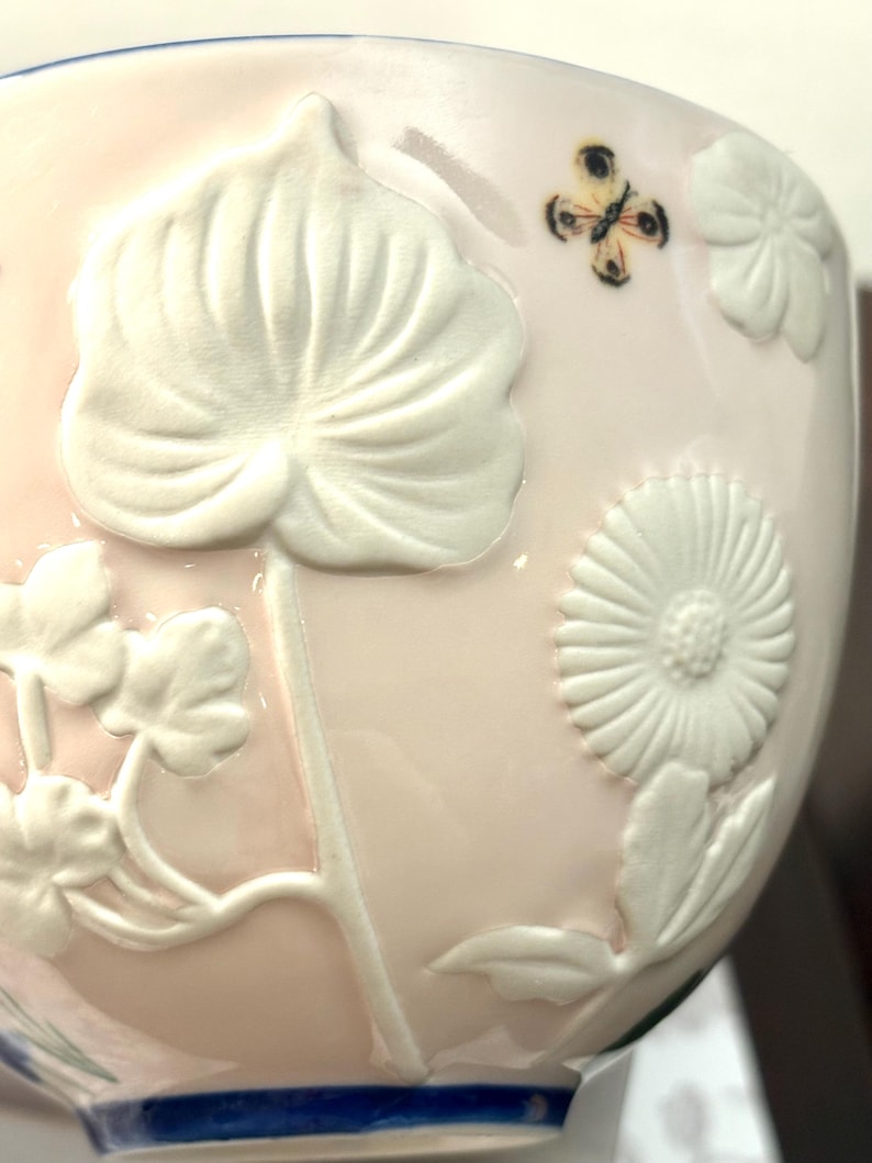 May include: A light pink ceramic bowl with white embossed floral and leaf designs. A small butterfly is painted on the side. The rim has a blue band. The bowl has a glossy finish.