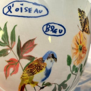 May include: A white ceramic mug with a hand-painted design featuring a blue and yellow bird perched on a branch, with orange flowers and a butterfly. The mug has the words "L'oiseau" and "Bleu" in blue text.