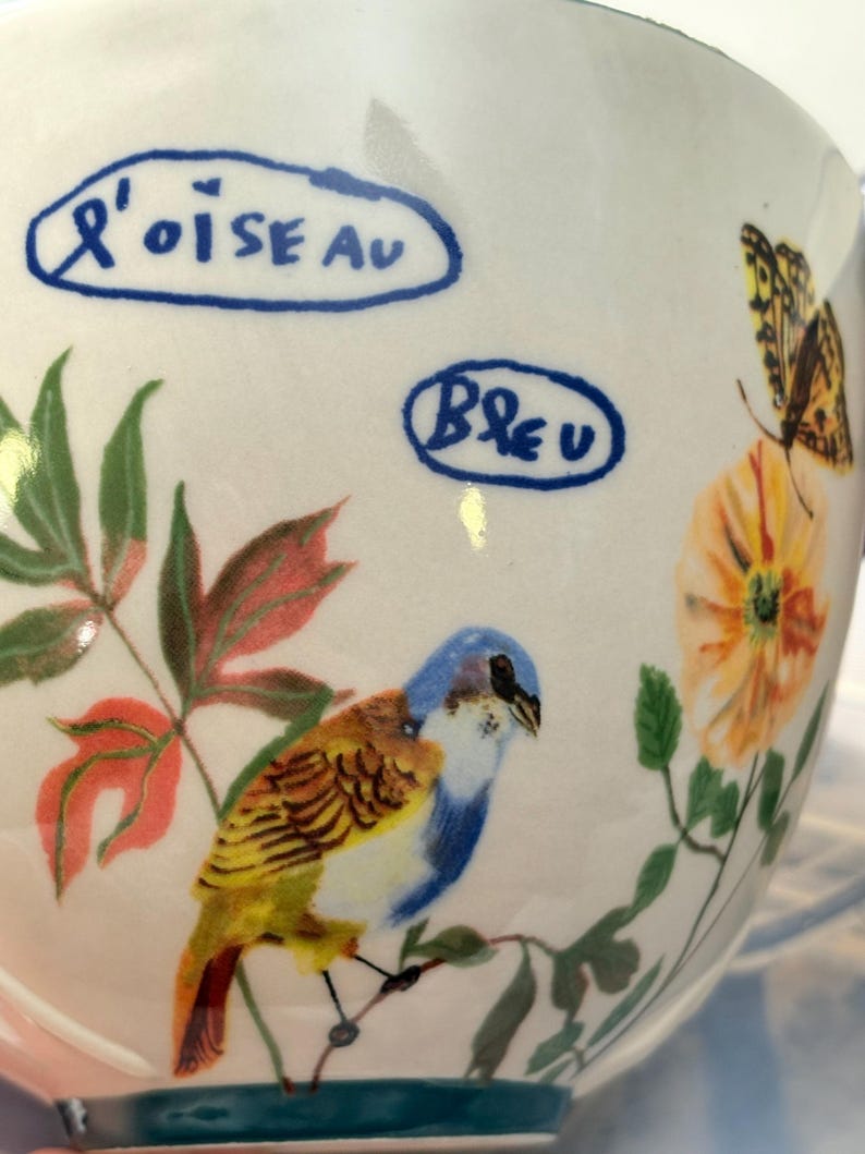 May include: A white ceramic mug with a hand-painted design featuring a blue and yellow bird perched on a branch, with orange flowers and a butterfly. The mug has the words "L'oiseau" and "Bleu" in blue text.