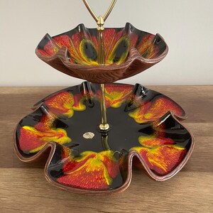 May include: Two-tiered serving dish with a gold metal stand. The dishes are black with red and yellow swirling patterns.