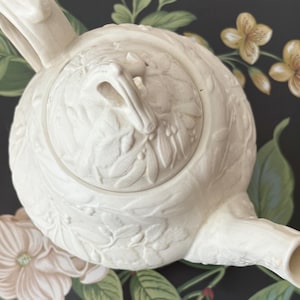 May include: Cream-colored ceramic teapot with a detailed floral relief pattern. The teapot features a curved spout, a handle, and a lid with a loop handle. The design includes leaves and berries, suggesting a botanical theme.