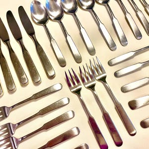 May include: An assortment of stainless steel silverware, including knives, spoons, and forks, arranged on a light-colored surface. The silverware has a simple, classic design, reflecting a vintage aesthetic.