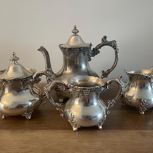 May include: A silver tea set with a teapot, creamer, sugar bowl, and a matching design. The set has an ornate design with floral accents.