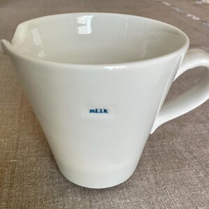 May include: White ceramic milk pitcher with a handle and a spout. The word "milk" is printed in blue on the front of the pitcher. The pitcher has a simple, modern design.
