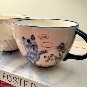 May include: A pink ceramic teacup with a blue handle and rim. The cup features a blue and white illustration of a cat, flowers, and a butterfly. The word "UNE" is written on the cup.