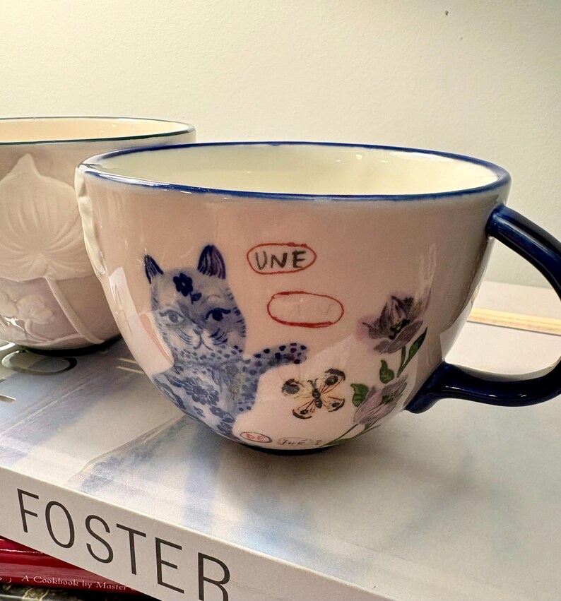 May include: A pink ceramic teacup with a blue handle and rim. The cup features a blue and white illustration of a cat, flowers, and a butterfly. The word "UNE" is written on the cup.