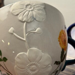 May include: A white ceramic mug with embossed floral designs and a painted yellow and orange flower. The mug has a dark green handle and rim, with the text "Catharine Bird" visible on the base.