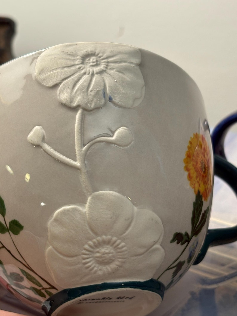May include: A white ceramic mug with embossed floral designs and a painted yellow and orange flower. The mug has a dark green handle and rim, with the text "Catharine Bird" visible on the base.