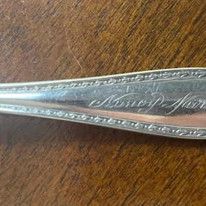 May include: Close-up of a silver spoon with an engraved inscription that reads "Nancy Marie".