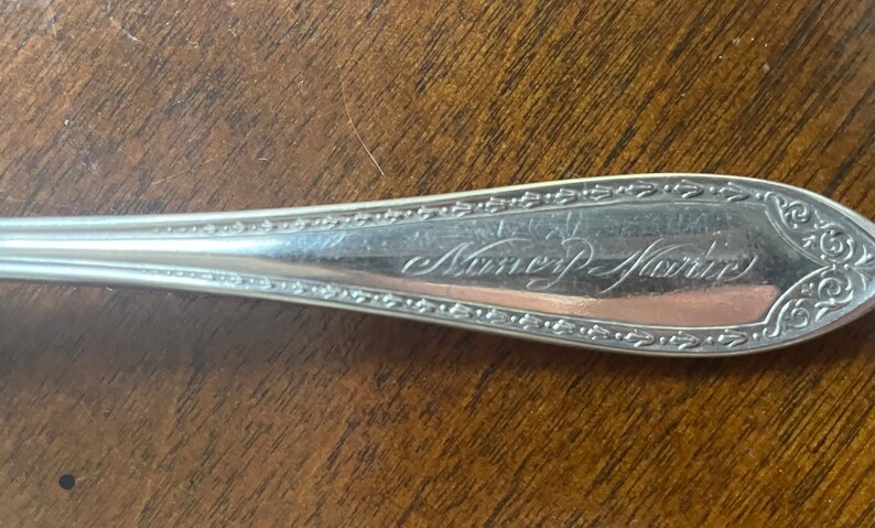May include: Close-up of a silver spoon with an engraved inscription that reads "Nancy Marie".