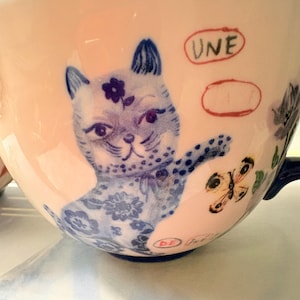 May include: A ceramic teacup with a light pink exterior, featuring a blue and white illustration of a cat with a flower on its head. The cup also has a butterfly and the word "UNE" in red.
