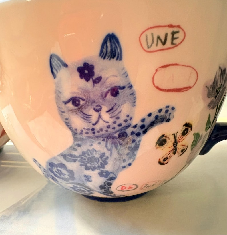 May include: A ceramic teacup with a light pink exterior, featuring a blue and white illustration of a cat with a flower on its head. The cup also has a butterfly and the word "UNE" in red.