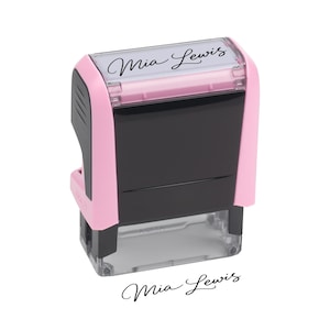 May include: A self-inking rubber stamp with a light pink and black plastic casing. The top display window features the name Mia Lewis in a black cursive script. Below the stamp, the same name is printed in black ink on a white background.