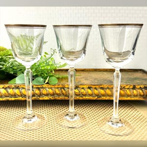 May include: Three clear glass wine glasses with a thin gold band around the rim. The glasses are on a gold and brown patterned surface.