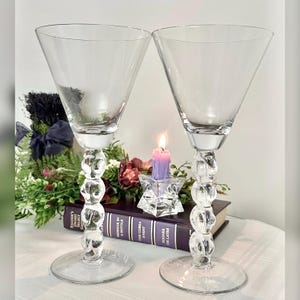 May include: Two clear martini glasses with a unique stem design featuring stacked, rounded shapes. The glasses sit on a clear, circular base. A lit purple candle and a book are in the background.