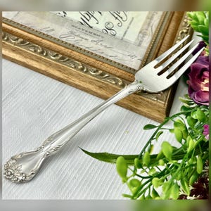 May include: A silver fork with ornate detailing on the handle, resting on a white surface. The fork is positioned in front of a framed document with gold trim. Green and purple floral accents are in the background.