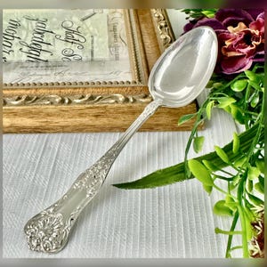 May include: A silver spoon with an ornate floral design on the handle and bowl. The spoon is set against a white tablecloth, with a wooden framed document in the background. The spoon's handle has a detailed, embossed pattern.
