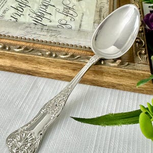 May include: A polished silver-toned metal spoon featuring an ornate, embossed handle with intricate scrollwork and a shell-like motif at the base. The spoon has a smooth, reflective oval bowl and a slender, decorative neck connecting to the handle.