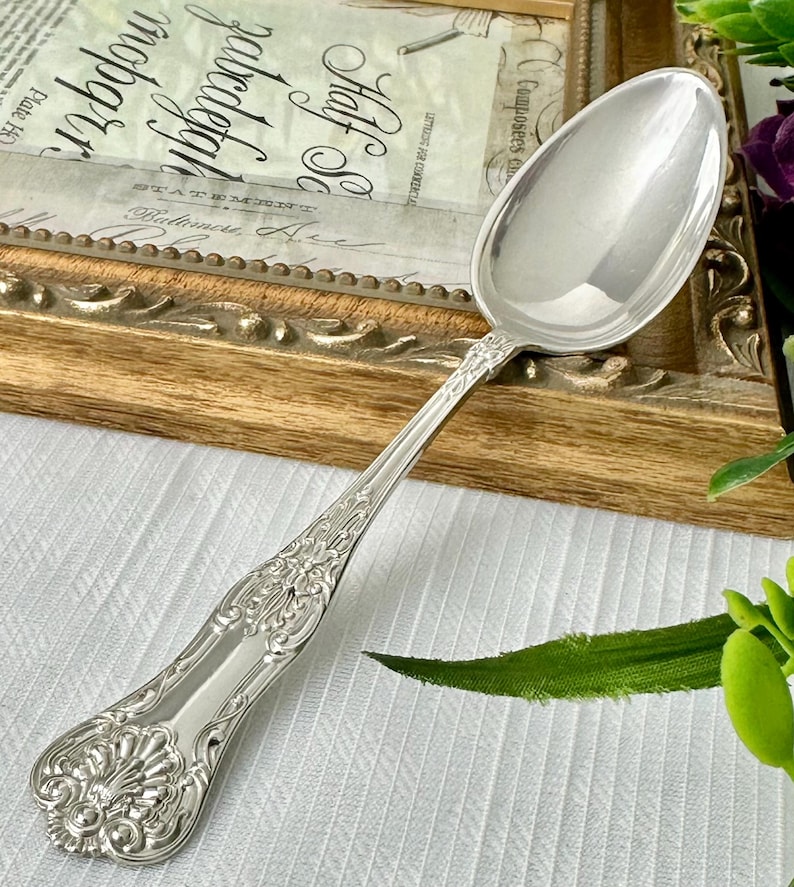 May include: A polished silver-toned metal spoon featuring an ornate, embossed handle with intricate scrollwork and a shell-like motif at the base. The spoon has a smooth, reflective oval bowl and a slender, decorative neck connecting to the handle.