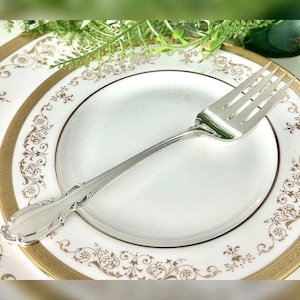 May include: A silver fork rests on a white plate with a gold floral border. The fork has a decorative handle and four tines. The plate is part of a set, with a matching design. The setting is elegant and suitable for a formal dining occasion.