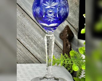 May include: A cobalt blue cut-to-clear crystal wine glass featuring a rounded bowl with intricate starburst and geometric patterns. The stem is clear, faceted, and textured, leading down to a circular, clear glass base with radial detailing.
