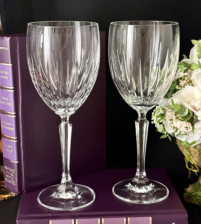 Pode incluir: Duas ta&ccedil;as de vinho de vidro transparente com um design de cristal cortado. As ta&ccedil;as est&atilde;o sobre um livro roxo com lombada dourada.