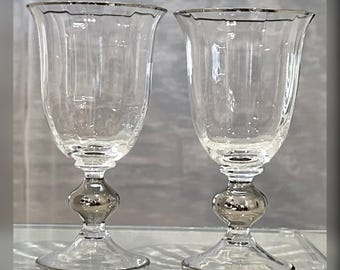 May include: Two clear glass goblets featuring a flared rim and a vertical ribbed pattern on the bowl. Each glass has a distinct, rounded, metallic silver-toned stem section that transitions into a clear, circular base with a silver rim.