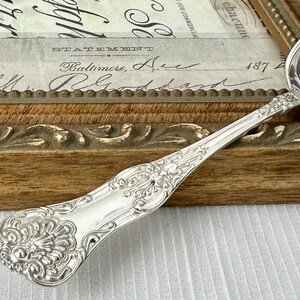 May include: A silver-toned metal spoon featuring an ornate, embossed handle with intricate scrollwork and a shell-like motif at the base. The handle tapers toward the bowl, showcasing detailed relief patterns that extend along the stem.