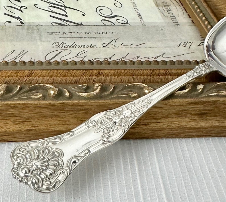 May include: A silver-toned metal spoon featuring an ornate, embossed handle with intricate scrollwork and a shell-like motif at the base. The handle tapers toward the bowl, showcasing detailed relief patterns that extend along the stem.