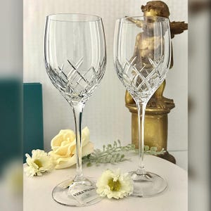 May include: Two clear crystal wine glasses with a cut-glass design. The glasses are on a white surface with flowers. The glasses have the text "Vera Wang" on the bottom.