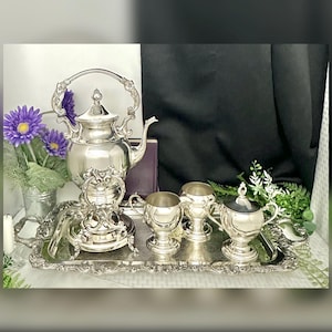 May include: A silver tea set with a teapot, sugar bowl, creamer, and cups on a decorative tray. The teapot has a curved handle and ornate detailing. Purple flowers and greenery are in the background.